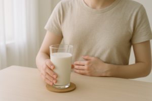 Persoon aan een lichte keukentafel met een glas melk en een hand op de buik